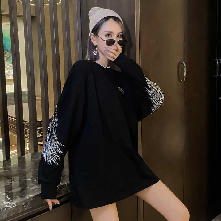 Person wearing a black oversized Maramalive™ Hot Diamond Mid-Length T-Shirt Women Long Sleeves with white graphics on the long sleeves, a grey beanie, sunglasses, and dangling earrings, partially covering their face with their hand while standing indoors.