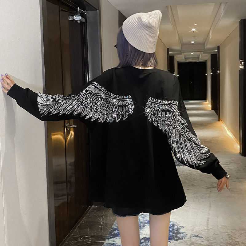 Person wearing a black, Maramalive™ Hot Diamond Mid-Length T-Shirt Women Long Sleeves with wing designs on the back, a beige beanie, and dark shorts, standing in a hallway with doors and overhead lighting.