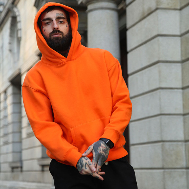 A person with tattoos and a beard, wearing an orange Maramalive™ Hoodie Hoodie over a crisp cotton shirt, poses in front of a stone building.