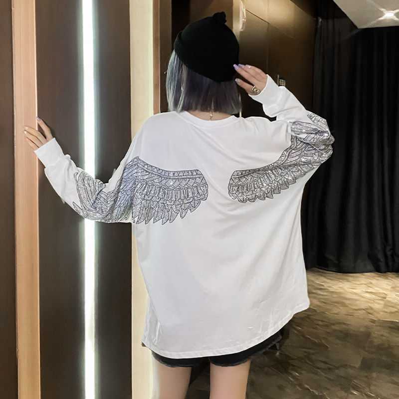 Person wearing a white, round-neck Maramalive™ Hot Diamond Mid-Length T-Shirt Women Long Sleeves with wing designs on the long sleeves, black hat, and shorts is standing with back to the camera, left hand touching a wall, in a dimly lit indoor setting.