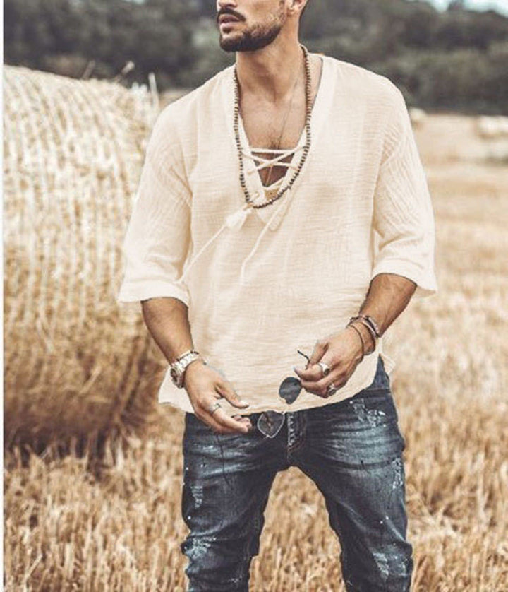 A man stands in a field wearing a white, Maramalive™ Men's Fashion Chest tie Mid Sleeve T Shirt with five-point sleeves and jeans. He has necklaces and bracelets, with a hay bale visible in the background.