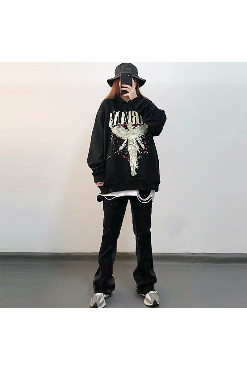 A person stands against a plain background wearing a black bucket hat, a black oversized punk style & hoodie sweatshirt: edgy look from Maramalive™ with a gothic fallen angel graphic, black pants, and sneakers.