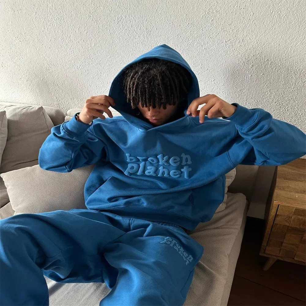 A street hipster sits on the couch, adjusting their hood. They sport a blue polyester hoodie and matching sweatpants, with "Letter Foam Printed Hoodie Punk Rock Casual Sweater" by Maramalive™ emblazoned on the front.