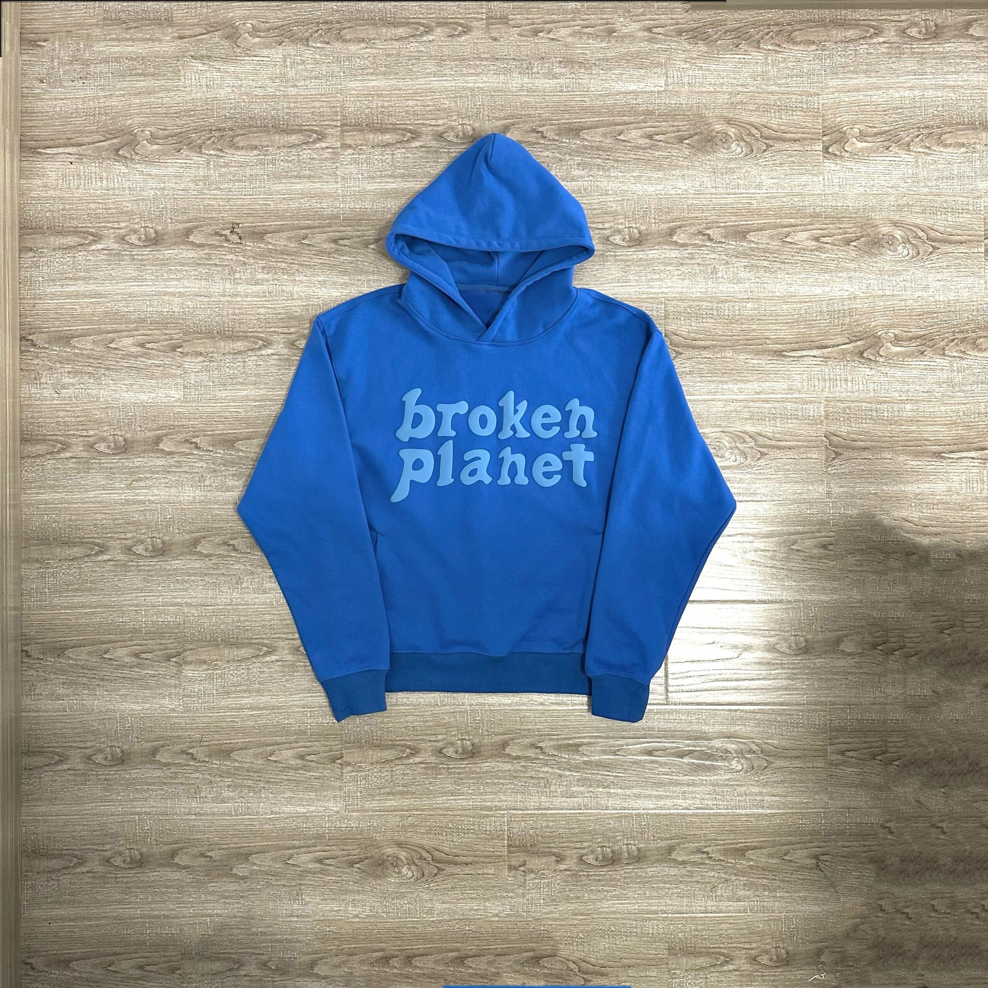 A blue polyester Maramalive™ Letter Foam Printed Hoodie Punk Rock Casual Sweater with the words "broken planet" written in white, laid flat on a wooden floor, perfect for any street hipster.