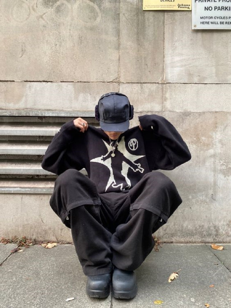Person wearing an oversized black Maramalive™ Hip-Hop Street Gothic Print Knitted Sweater with a Gothic style and a cap with ear flaps, sitting on the sidewalk and holding their sweater collar in front of a concrete wall with a metal grate and a parking sign.