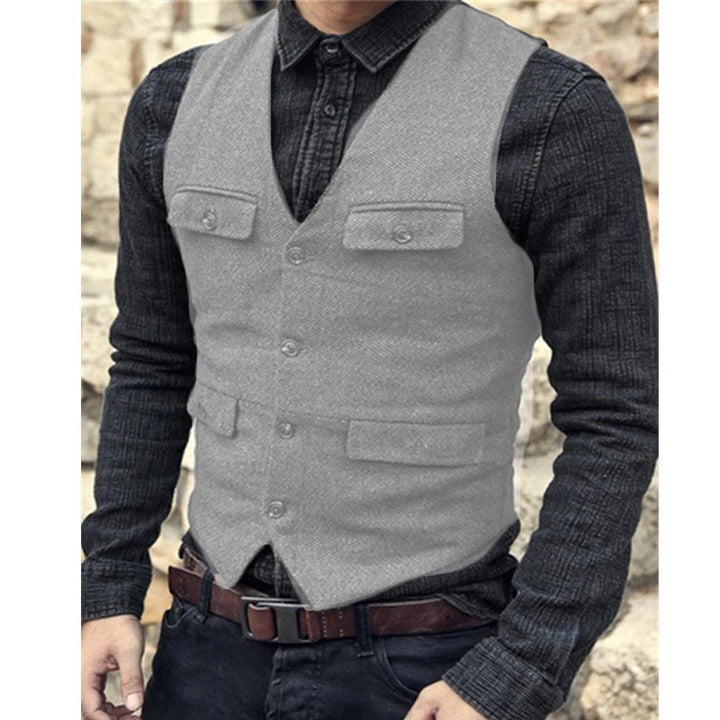 Person wearing a gray European And American Men's Vest Casual Solid Color Herringbone Vest by Maramalive™ over a dark denim shirt, with hands resting in pockets. The cotton blend vest, available in sizes from XS to XXXL, features four pockets – two on the chest and two near the waist. The backdrop appears to be a stone wall, adding to the British style aesthetic.