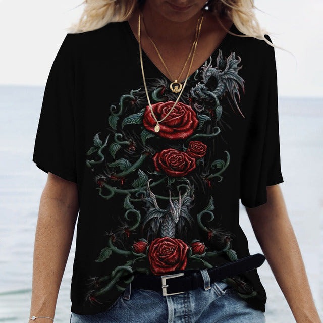 A person wearing a flattering fit, Ladies' Printed V-Neck Tee | Chic Women's Graphic Tees by Maramalive™ featuring a dragon and red rose design stands by the beach. The person is also wearing blue jeans and layered necklaces.