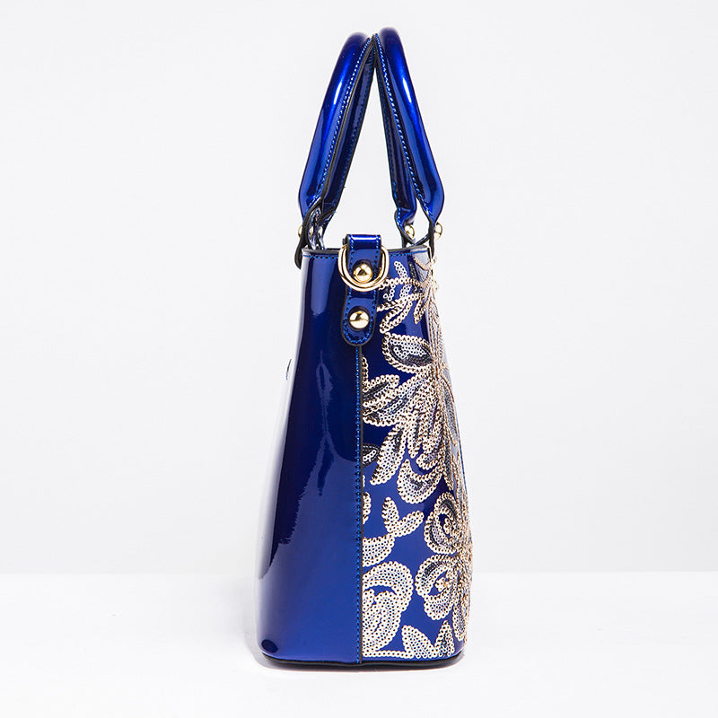 A shiny patent leather shoulder bag in blue with a floral design and zipper closure by Maramalive™.