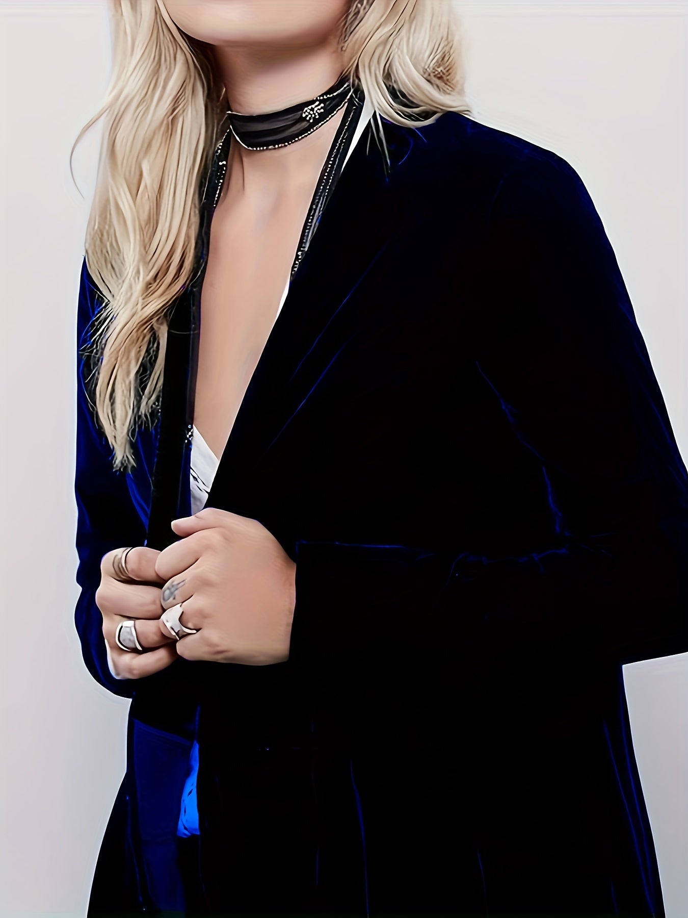 A person with long blonde hair wearing a Maramalive™ Women's Plus Solid Velvet Long Sleeve Open Front Ruffle Trim Longline Cardigan, white top, multiple rings, and two black chokers stands against a plain background. The outfit's medium stretch fabric ensures both comfort and style.