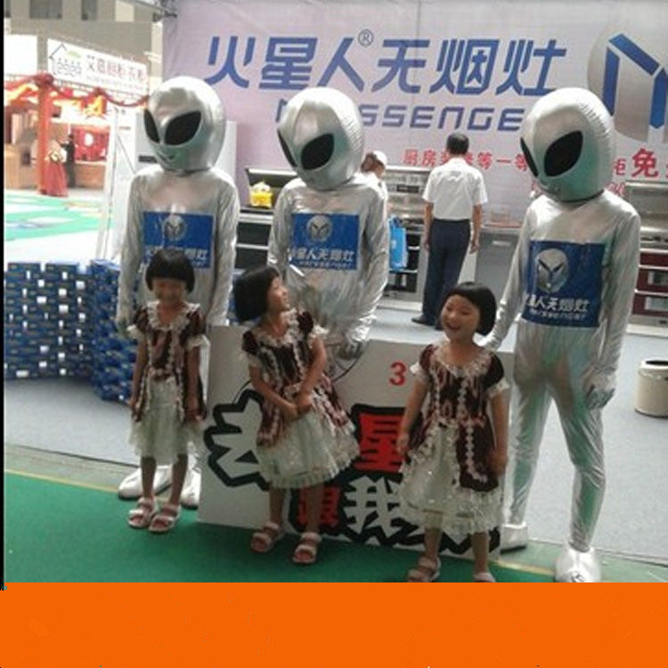 Three children stand smiling in front of a display with three people dressed in Maramalive™ Alien show costume Suits and helmets, at a themed sci-fi convention.
