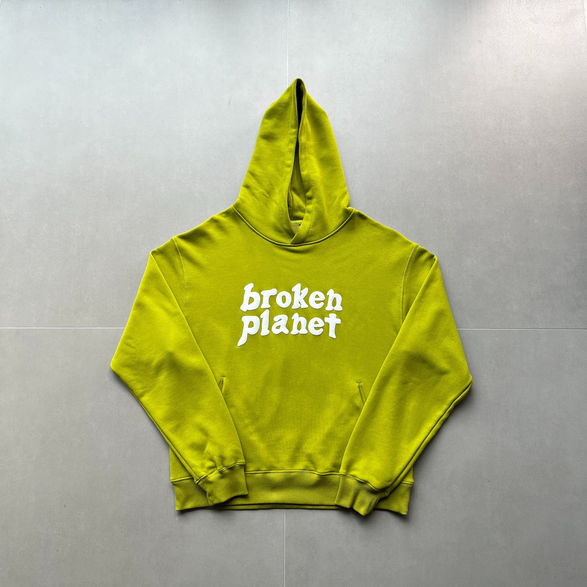 A lime green, polyester hoodie with "broken planet" printed in white text on the front, laid out flat on a grey tiled floor. This street hipster style Maramalive™ Letter Foam Printed Hoodie Punk Rock Casual Sweater is perfect for making a statement.
