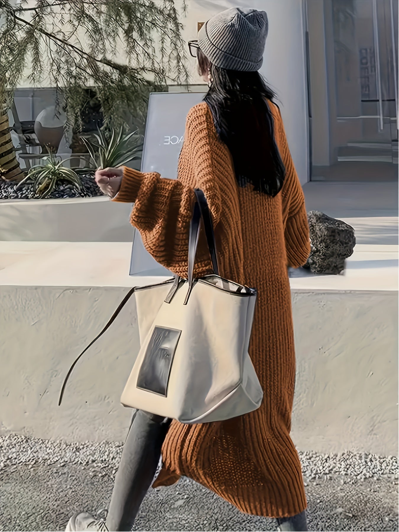 A person wearing a long orange Maramalive™ Plus Size Open Front Loose Knit Cardigan, Casual Long Sleeve Long Length Cardigan With Pocket, gray beanie, and glasses walks outdoors carrying a large cream-colored tote bag.