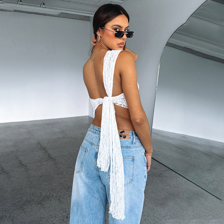 A person with long hair tied back is wearing a Maramalive™ Ins Lace Backless Top Summer Solid Color Waistless Asymmetrical Sloped Neck Vest Streetwear Womens Clothes and light blue jeans, standing in a spacious room with a large wall mirror. In their street hipster style, they are looking over their shoulder while wearing sunglasses.