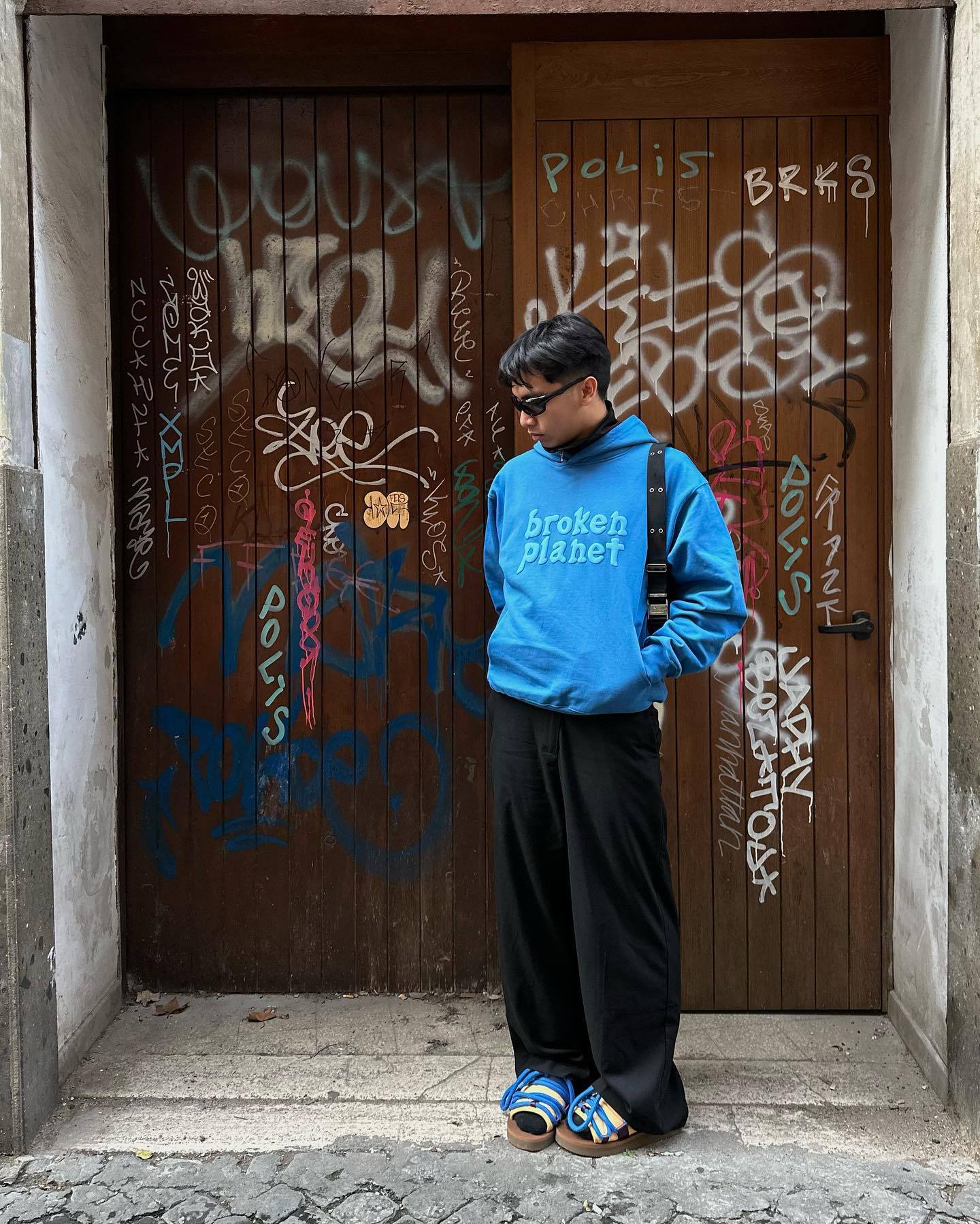 Person wearing a blue "broken planet" sweatshirt, black pants, and blue sandals, standing in front of a wooden door covered in graffiti. The street hipster effortlessly blends comfort and style, with their Maramalive™ Letter Foam Printed Hoodie Punk Rock Casual Sweater adding an extra layer of urban cool.