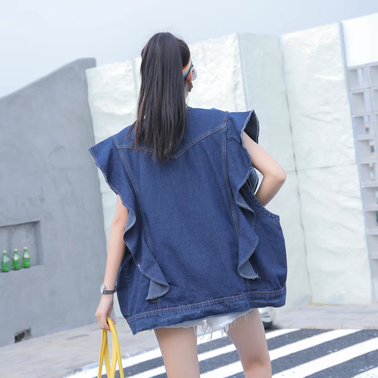 A person stands outdoors with their back to the camera, wearing a Maramalive™ Heavy Duty Diamond Studded Denim Vest With Wooden Ear Edge, holding a yellow bag, and sporting a headband.