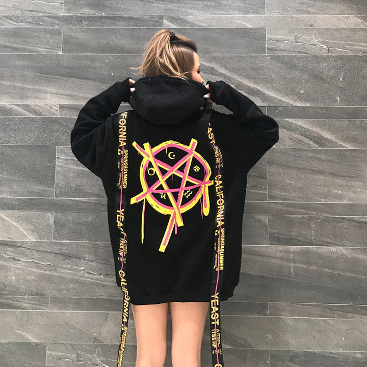 A person is wearing an oversized black MAGICIAN HOODIE by Maramalive™ with a neon pink and yellow geometric pattern and long straps that say "YEAST CALIFORNIA." The person has light-colored hair and is facing away from the camera, epitomizing street style.