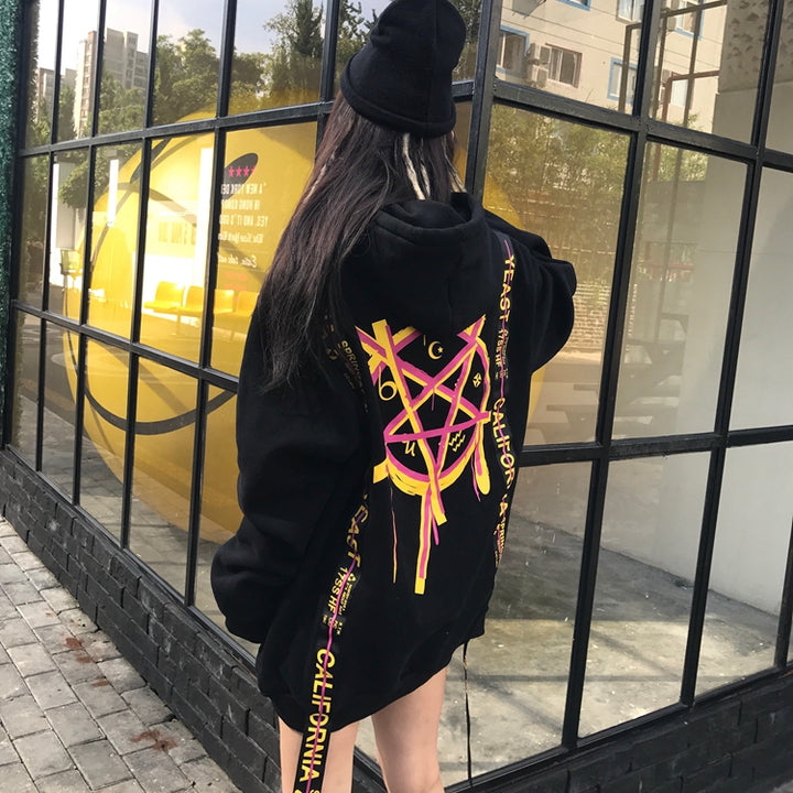 A person wearing a black Maramalive™ MAGICIAN HOODIE with a colorful geometric pattern on the back stands in front of a windowed building. The individual, exuding street style, sports a black beanie and has long hair.