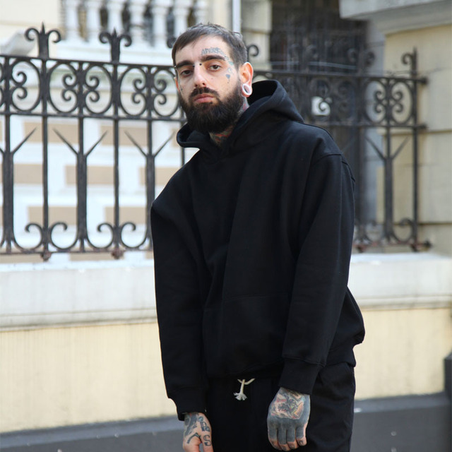 A person with face tattoos and stretched ear piercings is wearing a black Maramalive™ Hoodie Hoodie and pants, standing in front of a decorative iron fence and a building.