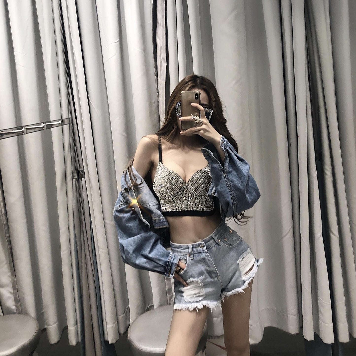 A woman stands in front of a mirror taking a selfie. She is wearing a Gypsophila shining studded waistcoat strapless tube top strap by Maramalive™, a denim jacket, and distressed denim shorts. Her long hair cascades over her shoulders as she holds a phone with her left hand, perfectly capturing the effortless style of her trendy ensemble.