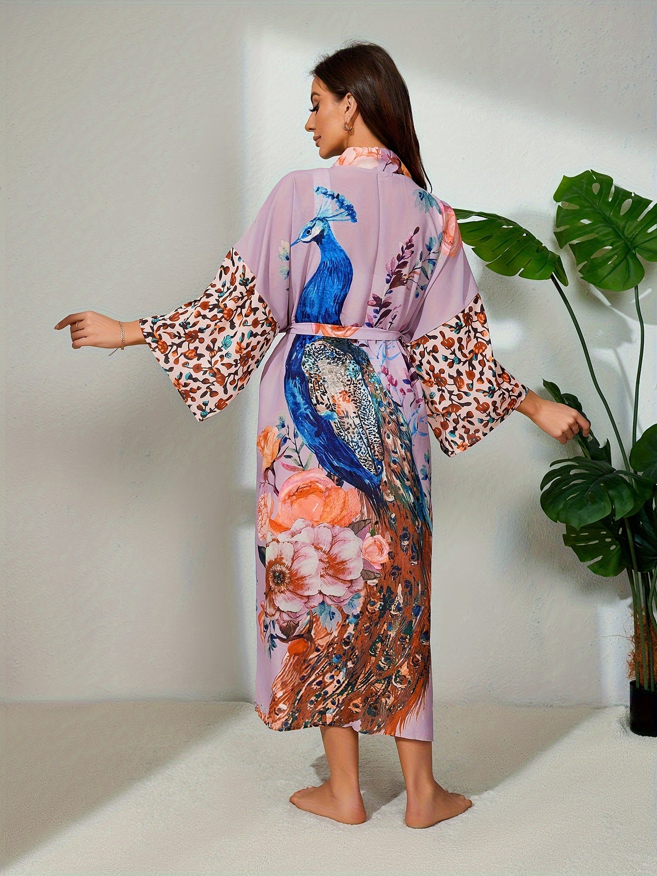 A woman stands with her back to the camera, wearing a Maramalive™ Bohemian Style Women's Peacock Print Beach Cover-Up With Belt, Long Sleeves Loose Fit Vacation Kimono. The boho tunic-style robe contrasts beautifully with the light-colored wall, as she stands near a tall potted plant.