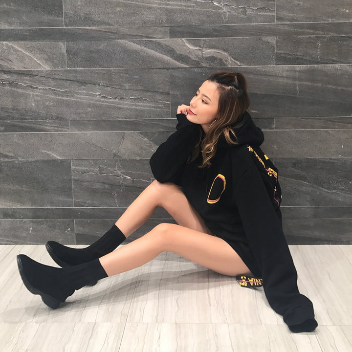 A person with long hair sits on a light floor against a dark, geometrically patterned wall, wearing a black Maramalive™ MAGICIAN HOODIE and black boots, resting their chin on their hand.