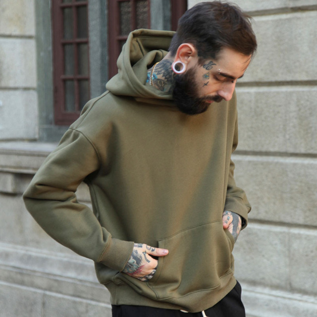 A bearded, tattooed man with ear gauges is wearing a green Maramalive™ Hoodie Hoodie, hands tucked in its pockets. He's standing outside near a building with a stone facade, looking down thoughtfully. The Maramalive™ Hoodie Hoodie complements his style as if he just checked the size chart before ditching his usual cotton shirt.