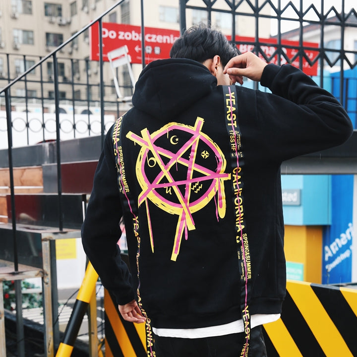 Person wearing a black Maramalive™ MAGICIAN HOODIE with a colorful geometric pattern on the back, standing outside on a city street near some black-and-yellow barriers.