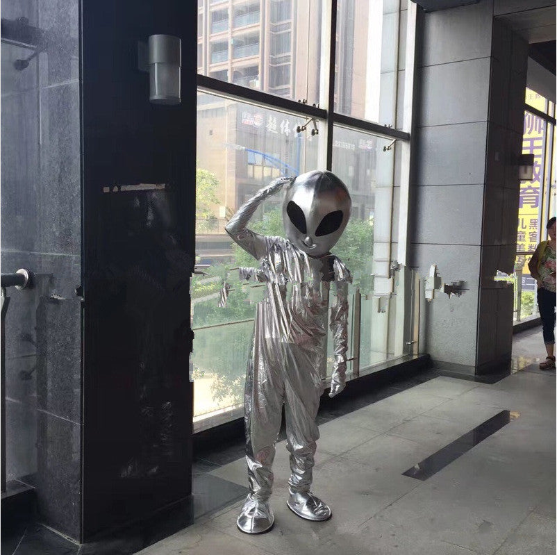 A person wearing a shiny silver Maramalive™ Alien show costume Suit with a large head and black eyes stands saluting inside a building with glass windows and tall structures outside, perfectly blending into the atmosphere of a lively sci-fi convention.