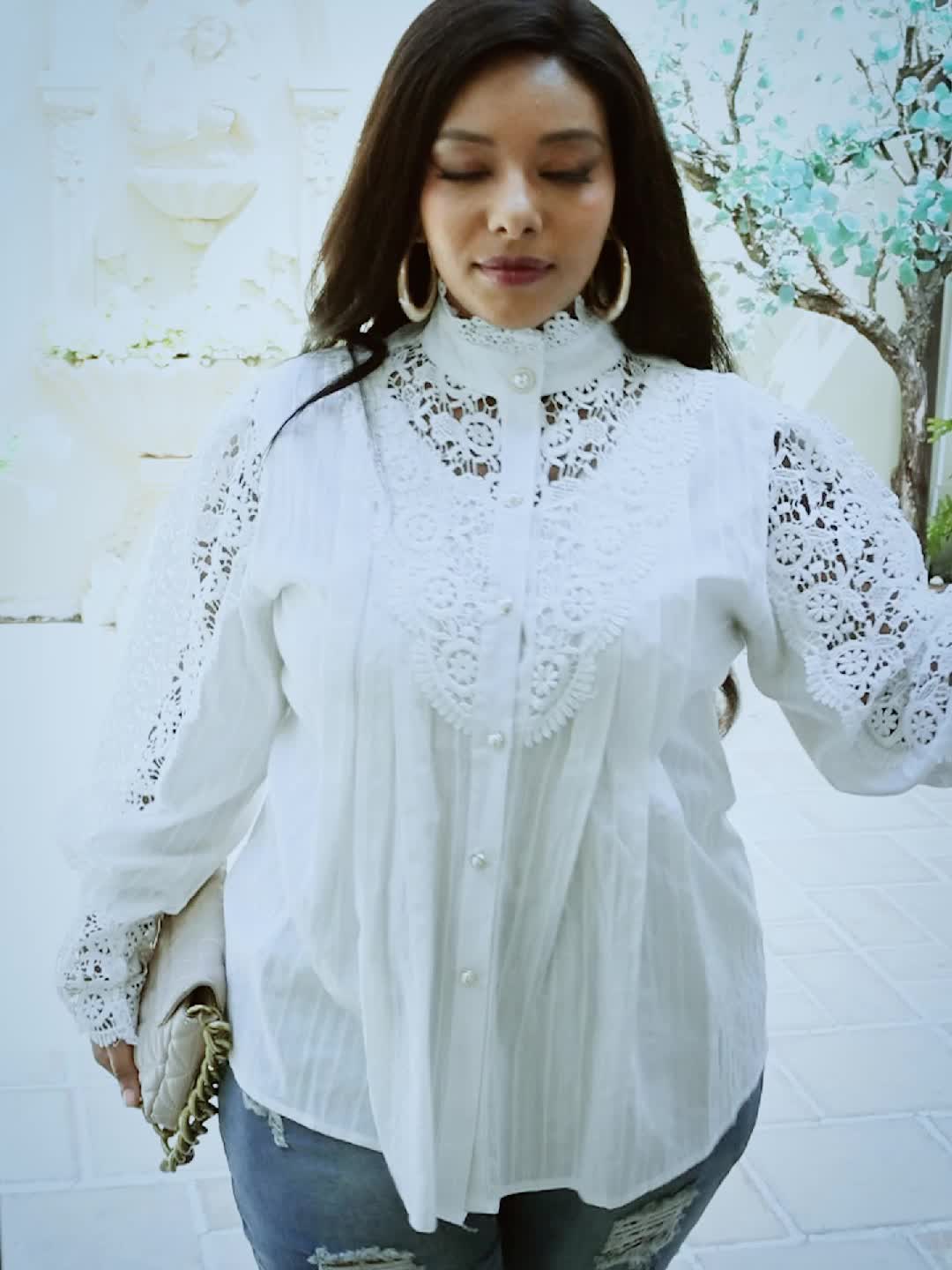 A woman wearing a white Maramalive™ Plus Size Elegant Blouse, Women's Plus Solid Contrast Lace Lantern Sleeve Button Up Mock Neck Shirt Top and holding a beige clutch stands outdoors against a light-colored background, with greenery visible in the distance. She is looking slightly downward.