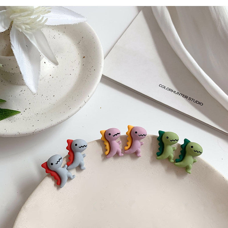 Women's Graceful And Fashionable Cute Dinosaur Stud Earrings by Maramalive™ on a table next to a cup of coffee.