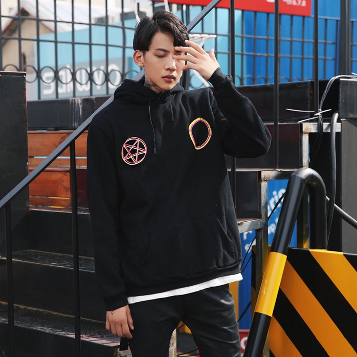 A man stands outdoors on stairs, wearing a black Maramalive™ MAGICIAN HOODIE with colorful circular and pentagram patches arranged in a geometric pattern, white shirt underneath, and black pants. Embracing street style, he touches his face with one hand, eyes closed.