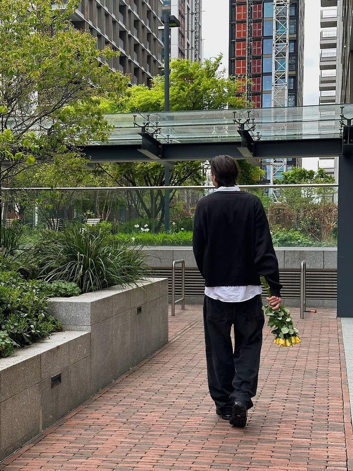 A person in dark clothing walks through an urban area, exuding a sense of gothic style. They hold a bouquet of flowers behind their back, contrasted against the geometric patterns of the tall buildings and greenery visible in the background, wearing a Hip-Hop Street Gothic Print Knitted Sweater from Maramalive™.