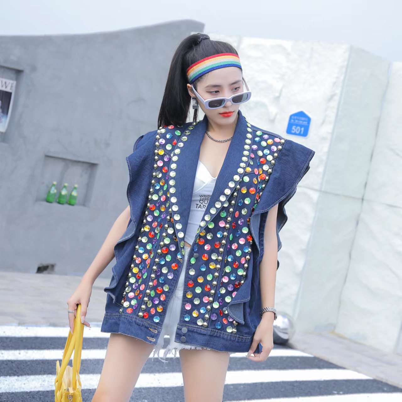 A woman wearing a colorful, Maramalive™ Heavy Duty Diamond Studded Denim Vest With Wooden Ear Edge with beaded sequins, white top, and mirrored sunglasses, holding a yellow bag and posing on a street. A rainbow headband adorns her high ponytail.