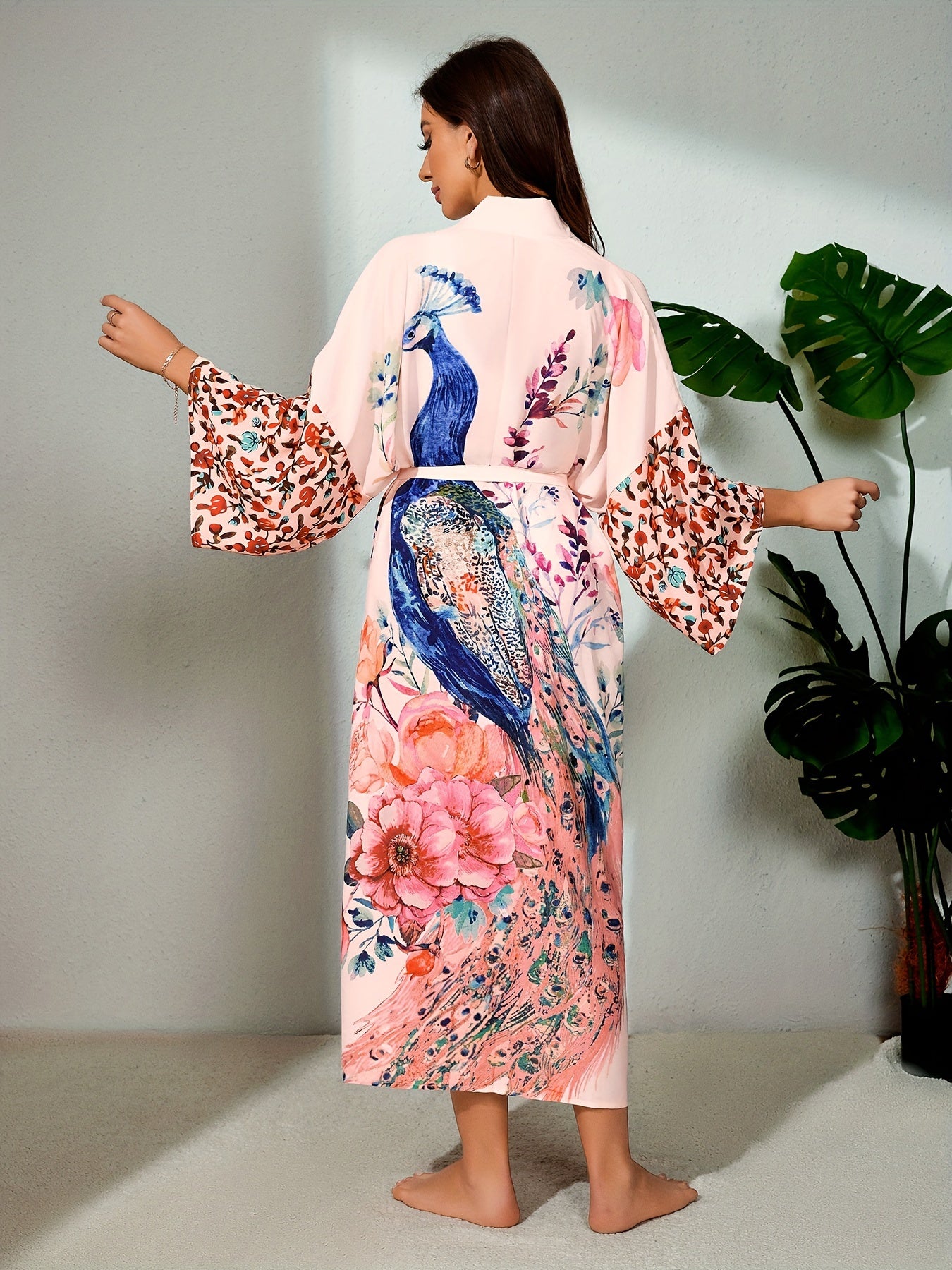 A woman in a long, colorful Maramalive™ Bohemian Style Women's Peacock Print Beach Cover-Up With Belt, Long Sleeves Loose Fit Vacation Kimono stands barefoot, facing away from the camera. A green potted plant is on the right side of the image, adding to the serene atmosphere.