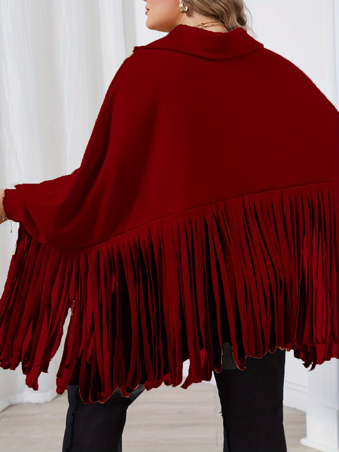 A person wearing a Maramalive™ Plus Size Trendy Top, Women's Plus Solid Batwing Sleeve Mock Neck Fringe Trim Cloak Top draped over their shoulders stands indoors with their back facing the camera.