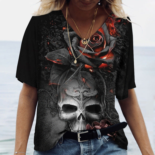 A person wearing a black Ladies' Printed V-Neck Tee from Maramalive™, featuring a detailed design of a skull and a rose, stands in front of a blurred background, possibly outdoors. The tee has short sleeves and offers a flattering fit.