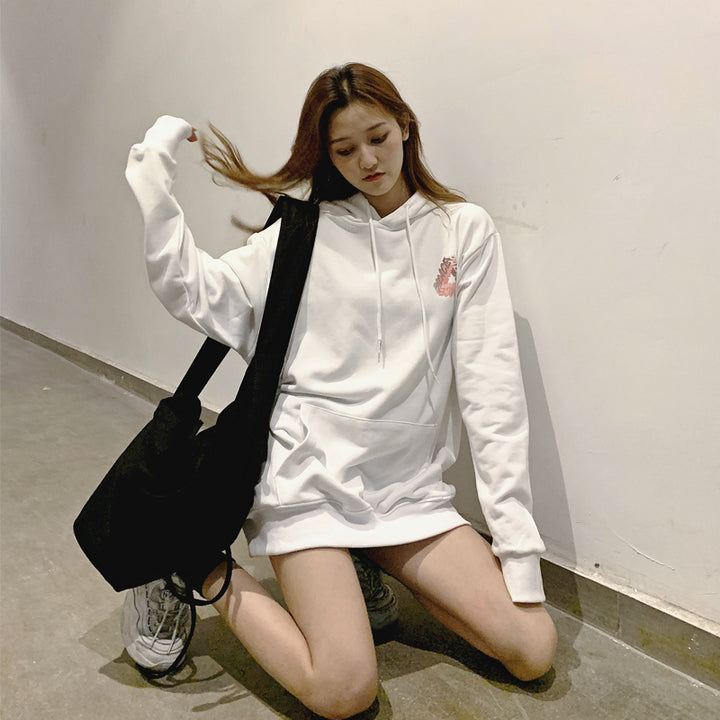 A person wearing a white Maramalive™ Phantom hoodie and sneakers, sitting on the floor indoors, holding a black bag and touching their hair. The scene captures the essence of youth fashion with clear hip hop influences, making it perfect for autumn and winter trends.