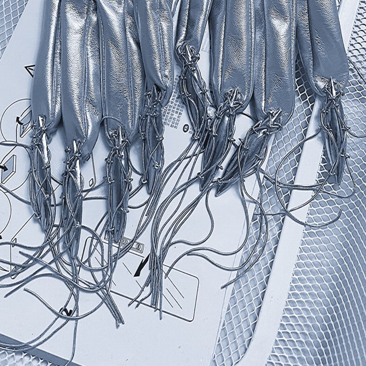 Several Maramalive™ Punk Futuristic Silver Metallic Performance Suits with long, thin cords at the fingertips are laid out on a white surface covered with printed diagrams and symbols, accompanied by delicate electroplated jewelry, all placed against a mesh backdrop.