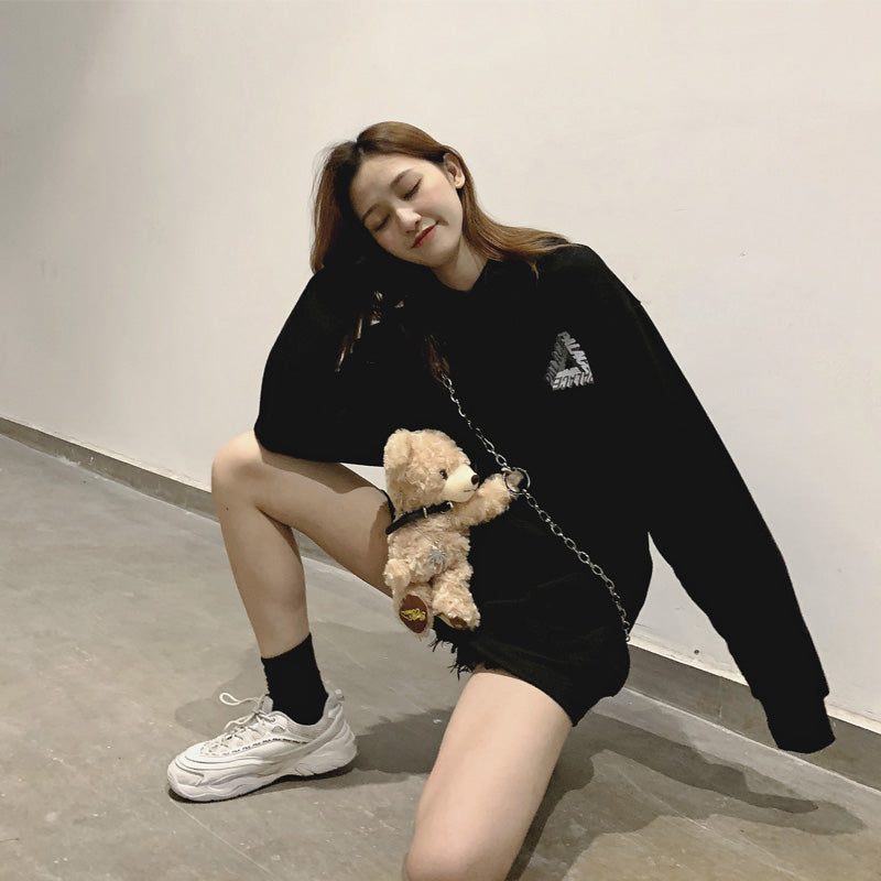 A person poses indoors in a black oversized Phantom hoodie by Maramalive™, black shorts, black socks, and white sneakers, embodying youth fashion with a hip hop twist. They hold a chain with a small teddy bear attached. The background is a plain light-colored wall and floor.