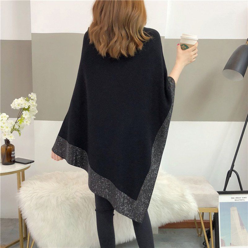 A person with brown hair, seen from behind, is standing indoors holding a coffee cup. They are wearing a Maramalive™ Outer Bat Sweater With Diamond Studded Black Sweater and black pants. The simple commuting style room includes a chair, table, and flowers.