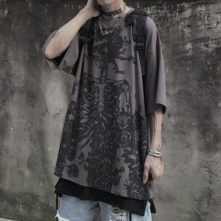 A person wearing a dark oversized graphic print tee, the Dark Hip Hop Tee - Perfect for Underground Rap Fans by Maramalive™, with a skeleton and floral design stands against a rough, textured wall. The individual, embodying streetwear fashion, also has tattoos, layered clothing, and is carrying a backpack.