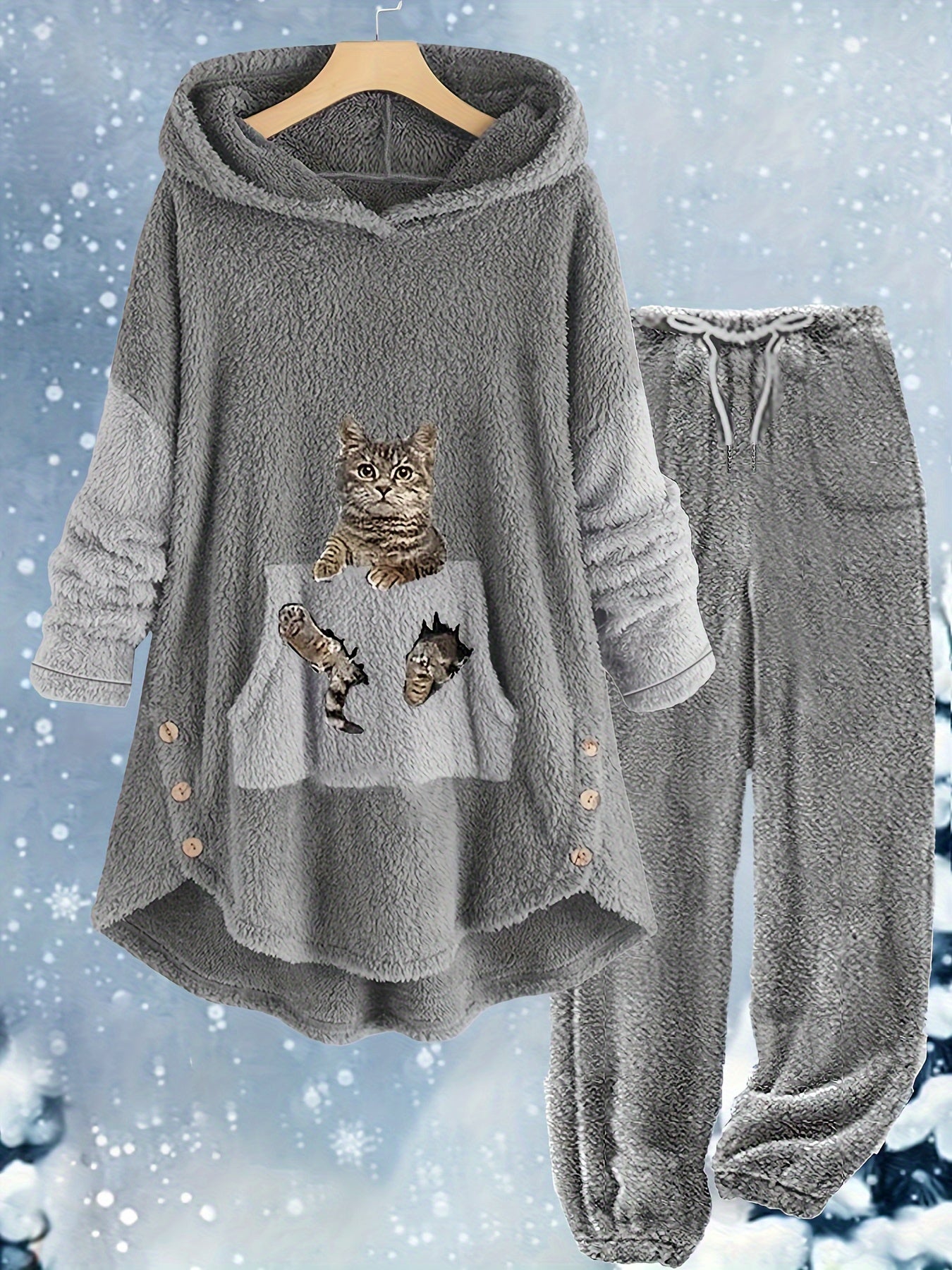 A grey polyester fleece loungewear set with a hooded top featuring a front pocket decorated with a cartoon image of a cat. The Maramalive™ Plus Size Casual Outfits Two Piece Set, Women's Plus Cat Print Fleece Long Sleeve Button Decor Hoodie & Pants Outfits 2 Piece Set is displayed on a hanger against a winter-themed background.