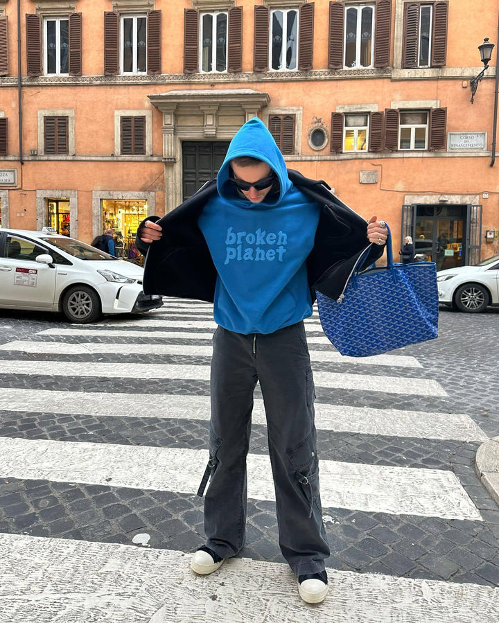 Person standing on a street crosswalk wearing a blue polyester Maramalive™ Letter Foam Printed Hoodie Punk Rock Casual Sweater, dark pants, white sneakers, a black jacket, sunglasses, and holding a blue tote bag. The street hipster look is effortlessly cool and casual.