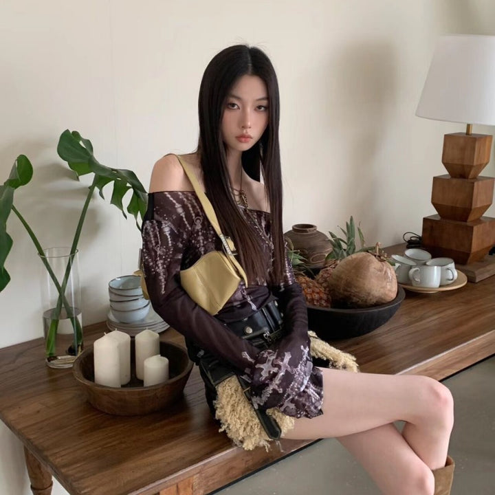 A young woman with long, straight hair sits on a wooden table next to a lamp, plants, candles, and various home decor items. She wears an urban style Maramalive™ Off-shoulder Printed Mesh Long Sleeve Bottoming Shirt Sun Protection Shirt and holds a small yellow bag.
