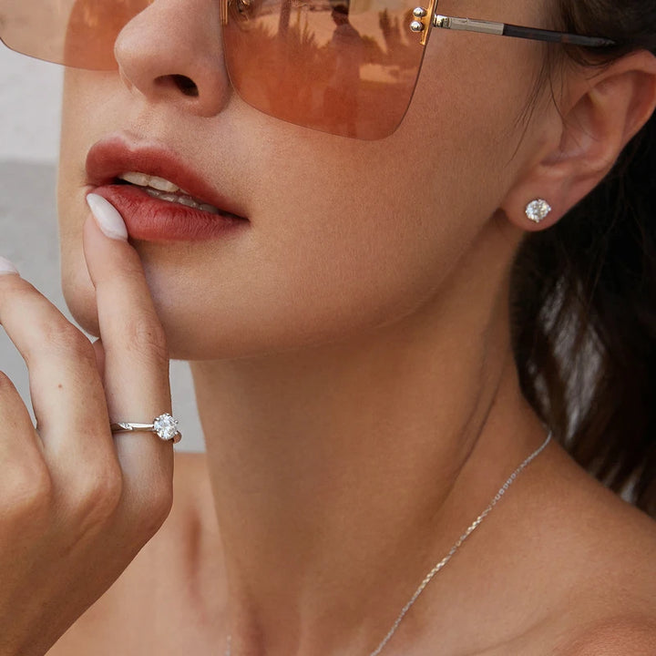 Woman wearing large sunglasses, diamond earrings, and a thin necklace with her hand touching her lips, featuring the stunning Maramalive™ Moissanite Ring 1ct Round Moissanite Diamond Solitaire Engagement 925 Sterling Silver Rings For Women.