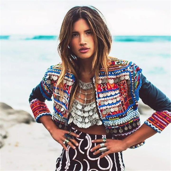 A person stands on a beach wearing a colorful, embroidered jacket, a metallic beaded top adorned with Maramalive™ Full Coins Tassel Chest Chain For Summer Beach Party Bohemian Gypsy Afghan Ethnic Slave Harness Underwear Bra Sexy Body Jewelry, and patterned pants.