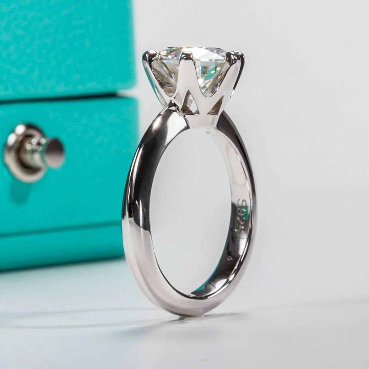 A Moissanite Solitaire Ring: Stunning Engagement Jewelry from Maramalive™ with a silver engagement ring featuring a solitaire design and a large moissanite center stone is displayed upright near a blue box.