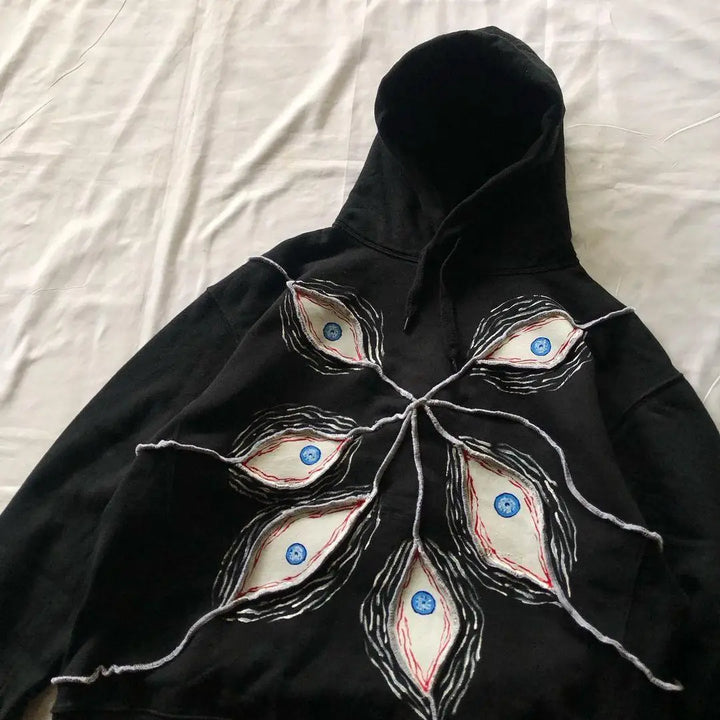 A Maramalive™ 2024 Fashion Goth Eye Mask Black Hooded Sweatshirt Retro Y2K Hip Hop Street Punk Casual Loose Jacket for Men and Women with intricate designs resembling eyes, connected by white thread-like lines, laid out flat on a light-colored surface. Perfect for Autumn/Winter casual pullovers.