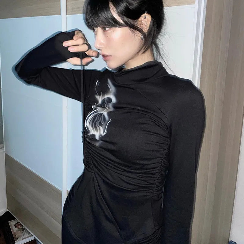 A person with dark hair, wearing a Maramalive™ Grunge Irregular Tops Dark Aesthetics Bandage T-shirt Mall Goth Y2k Tight Tops Alternative Streetwear Fashion Emo that exemplifies grunge style, is standing indoors and holding a string near their face. The room has light-colored walls and wooden furniture.