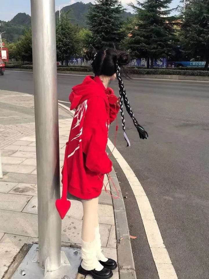 A person with long braided hair, wearing a vibrant red Maramalive™ Gothic Zip Up Hoodies Women Mall Goth Tops Streetwear Kawaii Hooded Sweatshirt 2022 Autumn Pullovers with a heart-shaped tail, stands by a pole on a sidewalk near a road with trees and parked cars in the background.